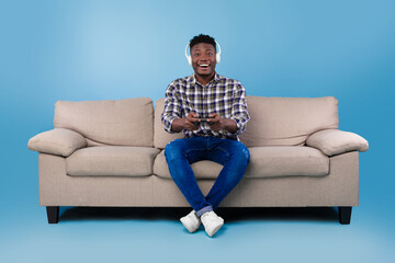 Wall Mural - Happy young black guy with joystick playing video game, wearing headset, sitting on couch over blue studio background