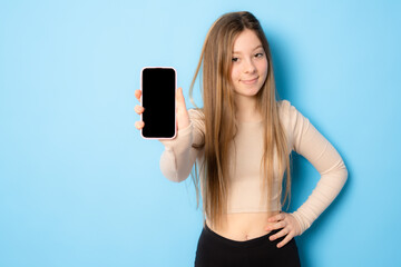 Stylish girl showing empty mobile phone screen and looking pleased, recommending application for smartphone, demonstrating shopping site over blue background
