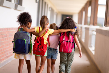 rear view of multiracial elementary schoolgirls with backpacks and arm around walking in corridor