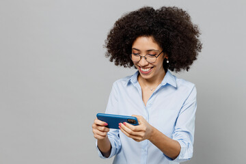 Poster - Young fun employee business corporate lawyer woman of African American ethnicity in classic shirt work in office use play racing app on mobile cell phone gadget smartphone isolated on grey background.