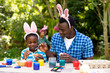 African american boy and father painting easter eggs while family in background