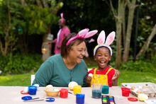 Cheerful African American Girl And Grandmother Wearing Bunny Ears While Painting Eggs On Easter Day