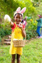 Portrait Of Happy African American Girl In Bunny Ears Showing Easter Egg While Family In Backyard