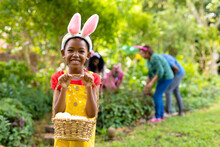Happy African American Girl In Bunny Ears Carrying Basket Of Easter Eggs While Family In Background