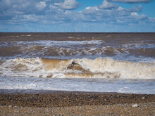 Rolling Waves Crash Onto The Beach With The White Wind Turbines Of A Massive Wind Farm On The Horizon, Blakeney Point, Cley Next The Sea, Norfolk, UK