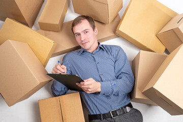 Courier job. Man is lying on the floor in the middle of cardboard boxes. Delivery man with notebook. A lot of orders for delivery. Great demand for courier services. Courier in a plaid shirt