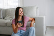 © Evrymmnt - Portrait of young brunette woman wearing mom jeans sitting on the floor leaning on the couch texting and smiling. Joyful female having fun video phone call. Background, copy space, close up.