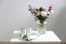 Bouquet Of Hackelia Velutina, Purple And White Roses, Small Tea Roses, Matthiola Incana And Blue Iris In Glass Vase Is On White Coffee Table With Two Tall Cups For Tea. Grey Wall