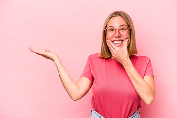 Wall Mural - Young caucasian woman isolated on pink background holds copy space on a palm, keep hand over cheek. Amazed and delighted.