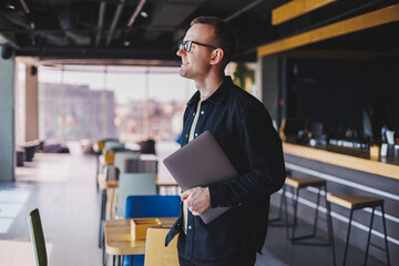 Male entrepreneur in black shirt and glasses standing in office with laptop in hand, successful corporate boss feeling good from rich lifestyle