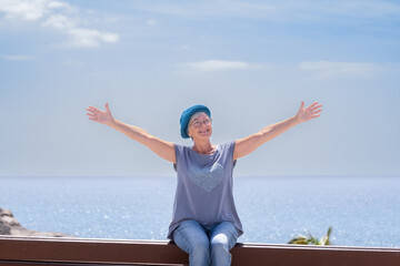 Sticker - Smiling senior woman with blue cap sitting on bench close to the sea with open arms. Elderly lady relaxing enjoying vacation or retirement under the sun. Horizon over water