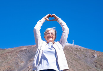 Feeling of freedom for a smiling senior white haired woman standing on mountain background making heart shape with her hands. Elderly happy woman enjoying travel and vacation