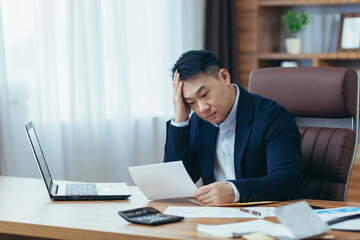 Young asian male accountant, office worker working with documents, tired, sitting at desk in office