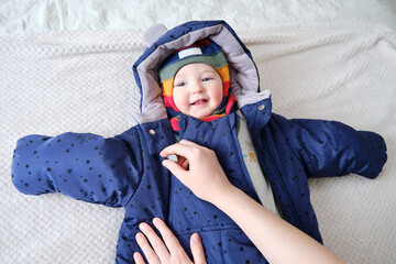 A mother woman puts on a warm jacket for a baby boy. Mom dresses a happy child in winter clothes on the bed.