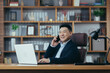 © Liubomir - Portrait of young handsome asian man businessman, lawyer, director in black suit. He talks on the phone, smiles, sits in a chair at a desk in the office, works with a laptop and documents.