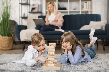 Little children sit on warm floor in living room play construct with wooden building blocks bricks together make a tower, young mom rest on sofa in background, Caucasian family relax at home