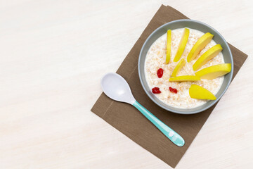 Funny cute kids childrens babys breakfast lunch oatmeal porridge in bowl look like hedgehog decoration with apple, dried cherry berry fruits, food art on white wooden table with copy space