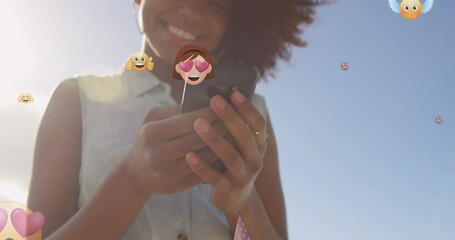 Sticker - Animation of emoticons over midsection of biracial woman using smartphone outdoors