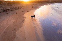 Aerial View Of A Two Surfers Walking Along A Beach Towards The Sunset