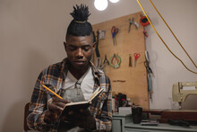 African American Young Craftsman With Dreadlocks Writing In Book While Working In Leather Workshop