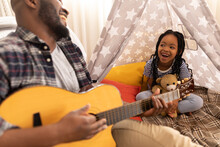 Cheerful African American Daughter And Father Sitting With Teddy Bear And Guitar In Tent At Home