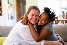 Portrait Of Happy Caucasian Mature Mother And African American Girl Embracing While Sitting On Sofa