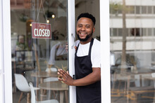 Smiling Portrait Of African American Male Barista Standing At Coffee Shop Doorway With Open Sign