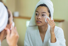 Biracial Young Woman Wearing Bathrobe Applying Cream On Face In Front Of Mirror In Bathroom