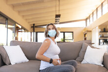 Portrait of caucasian pregnant woman wearing a face mask touching her belly sitting on the couch