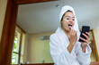 © Wavebreak Media - Biracial young woman in towel and bathrobe using smart phone while brushing teeth in bathroom