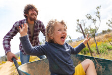Happy Young Man Pushing Excited Son Shouting While Sitting In Wheelbarrow At Farm