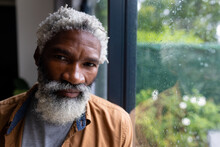 Portrait Of Confident African American Senior Man With White Beard Leaning On Window Glass At Home