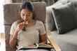 © Prathankarnpap - Satisfied asian woman drinking coffee and reading book on armchair at home.