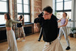 © pressmaster - Flexible teenage guy in black sweatshirt and white pants training in dance hall or modern studio against other performers exercising