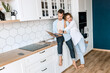 © shunevich - Lovely family couple are standing at the kitchen at home, doing online shopping or chatting with friends. Joyful stylish husband and wife using laptop, watching funny videos or photos, smiling