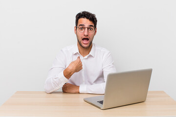 Wall Mural - hispanic handsome man looking shocked and surprised with mouth wide open, pointing to self. businessman and desk concept