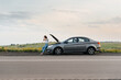 © Andrii - A young girl stands near a broken car in the middle of the highway during sunset and tries to call for help on the phone and start the car. Waiting for help. Car service. Car breakdown on the road.