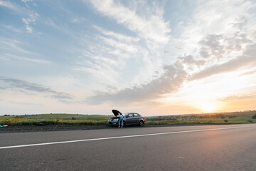 Wall Mural - A young girl stands near a broken-down car in the middle of the highway during sunset and tries to call for help on the phone. Waiting for help. Car service. Car breakdown on road.