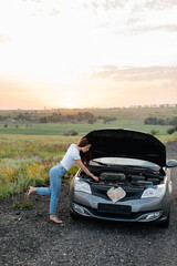 Wall Mural - A young girl stand near a broken car in the middle of the highway during sunset and tries to repair it. Troubleshooting the problem. Car service. Car breakdown on the road.