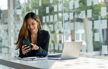 Young beautiful asian woman using smartphone and working with laptop while sitting at office desk, working from home concept.