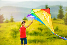 School-age Boy Run Huge Colored Rainbow Kite With Long Tail At Sunset Or Sunrise In Summertime. Mountain Landscape With Coniferous Trees On Horizon.