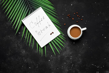 Wall Mural - Coffee break. A cup of coffee with an open notepad and decorative palm leaves on a black stone background. Top view.