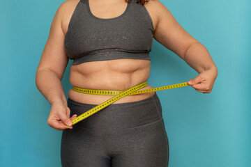 Wall Mural - Cropped photo of fat plump overweight woman standing in grey sports bra and leggings, showing excess naked belly, measuring waist, holding tape on blue background. Dieting, unhealthy food, obesity.