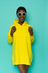Wall Mural - Smiling young black woman in sunglasses and vibrant yellow oversized hooded sweatshirt.
