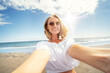 Young beautiful blonde woman in sunglasses smiling to the camera while taking selfie on the sandy beach. Girl enjoying summer vacation concept