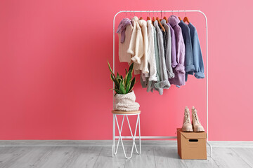 Rack with stylish warm sweaters, houseplant and boots near pink wall