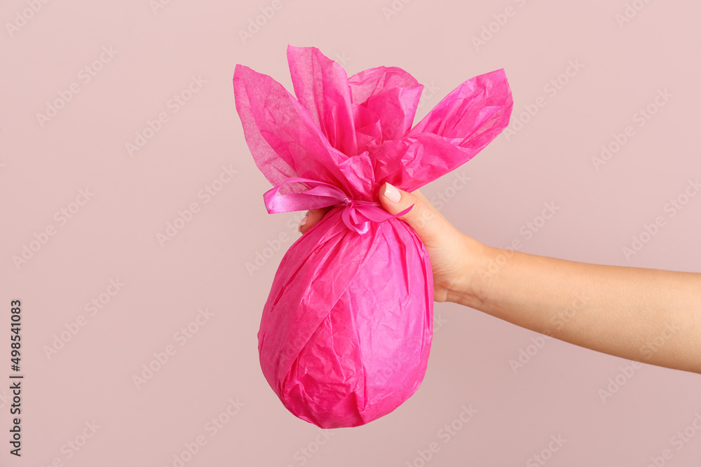 Female hand with chocolate Easter egg wrapped in pink paper on color background