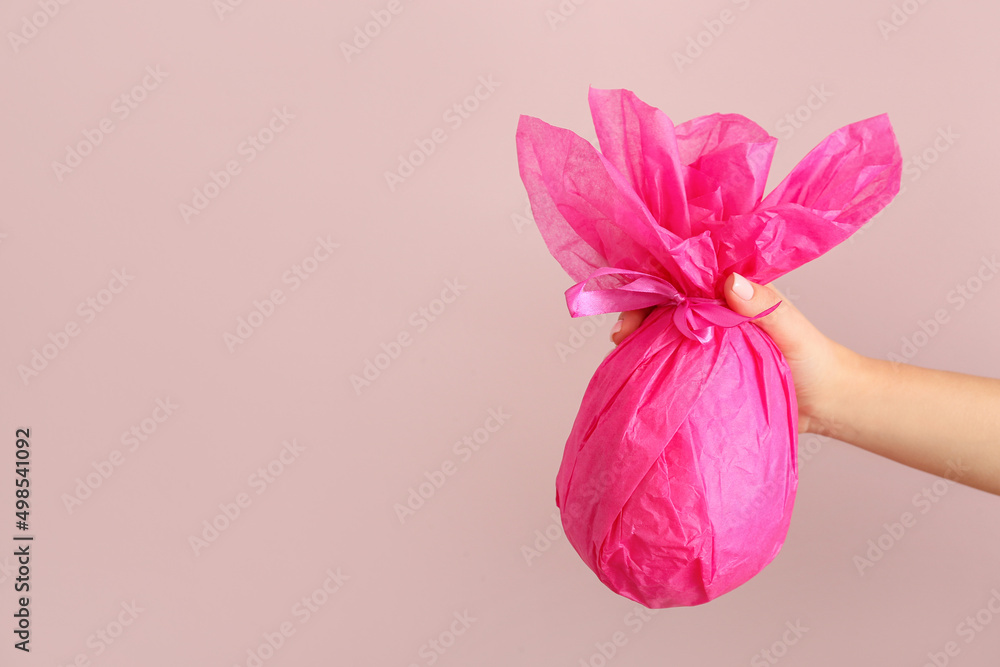 Female hand with chocolate Easter egg wrapped in pink paper on color background