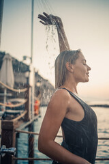 Wall Mural - A beautiful young attractive woman in a black swimsuit with a beautiful figure stands under a summer shower on the pier by the sea. She is enjoying her vacation. Selective focus