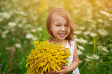 Funny Red-haired Girl Laughs Merrily Looking Into The Camera With A Bouquet Of Yellow Flowers In Her Hands In A Field At Sunset. Sincere Fervent Children's Laughter. Meadow With Achillea Millefolium